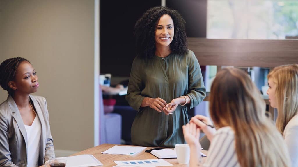 Mulher negra liderando reunião em ambiente de trabalho, simbolizando liderança feminina e gestão humanizada, com foco na colaboração, comunicação empática e inclusão entre a equipe presente.