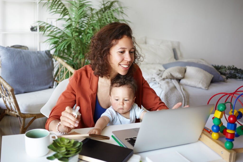 Mulher trabalhando em um notebook com um bebê no colo dentro de casa, ilustrando a realidade de negócios e maternidade. A imagem representa o empreendedorismo materno, a busca por equilíbrio entre carreira e família e a criação de negócios inspirados pela maternidade.