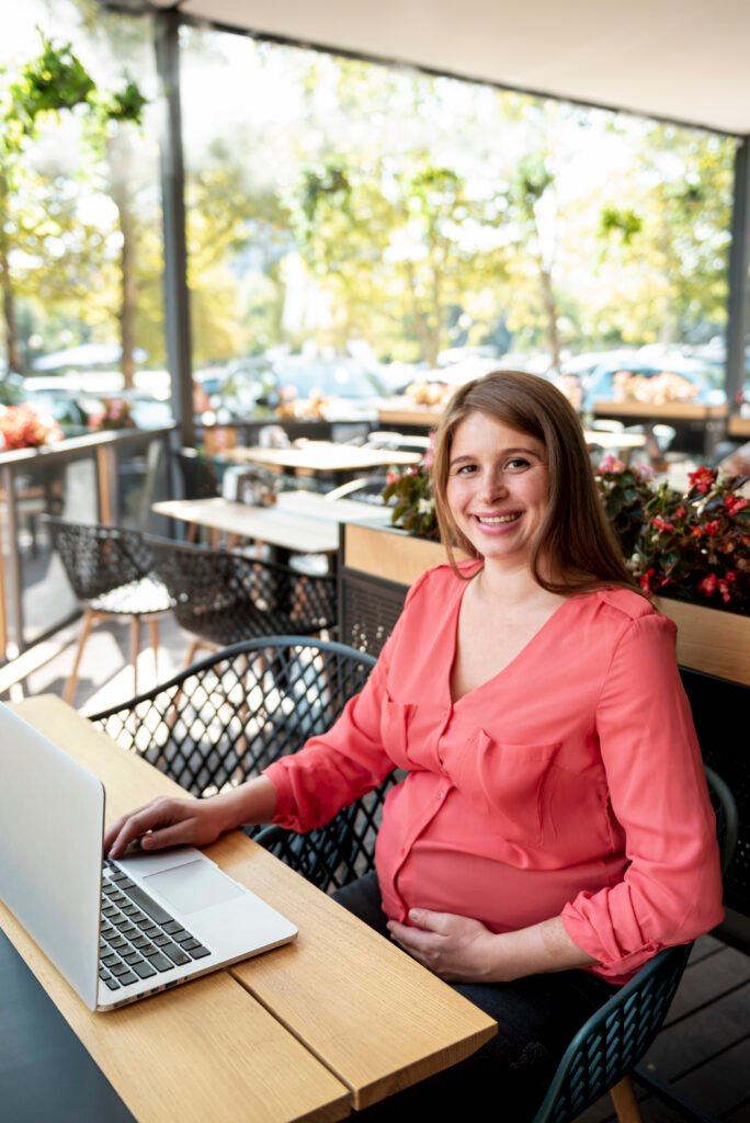 Mulher grávida utilizando um notebook em uma cafeteria ao ar livre, representando os negócios e maternidade e a preparação para conciliar maternidade, carreira e empreendedorismo. A imagem simboliza planejamento, autonomia profissional e a criação de negócios inspirados pelas transformações da maternidade.