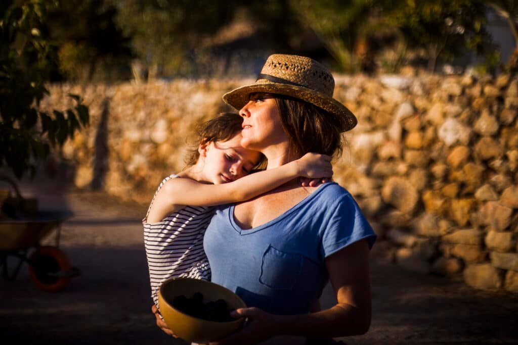 Mãe segura uma criança no colo em um ambiente ao ar livre, iluminado pela luz do fim da tarde. Enquanto carrega uma tigela com alimentos, a criança descansa a cabeça em seu ombro em um gesto de carinho. A imagem transmite acolhimento, cuidado, força e a conexão entre mãe e filho, representando os desafios e a dedicação da maternidade solo.
